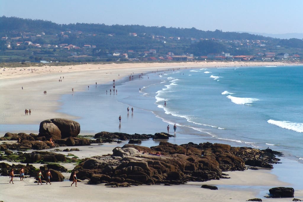 The long sweep of Praia da Lanzada with rocks in the foreground and the chapel-side village beyond